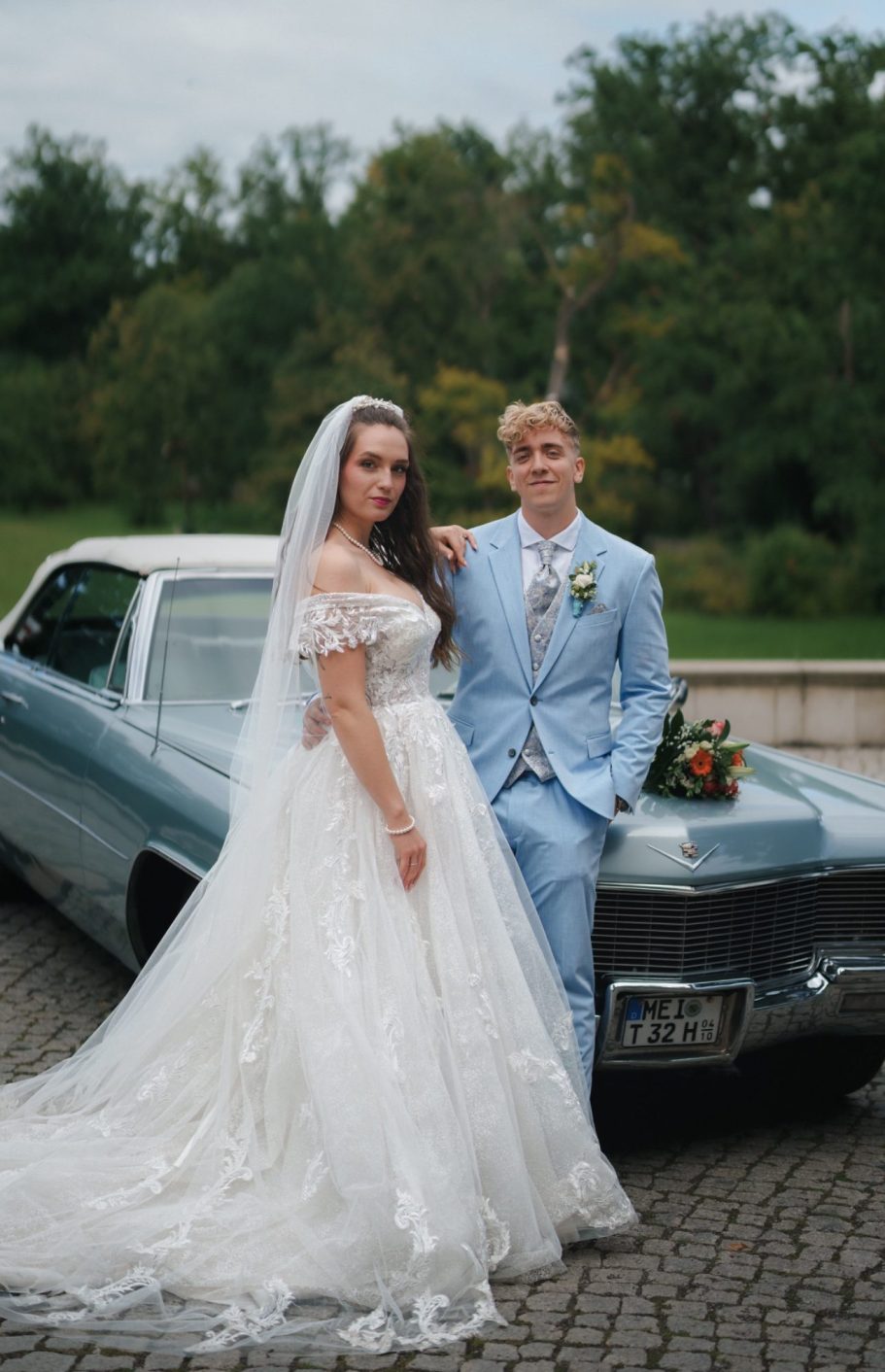 Brautpaar an Oldtimer Braut und Bräutigam posieren vor einem klassischen Auto in einer grünen Umgebung.
